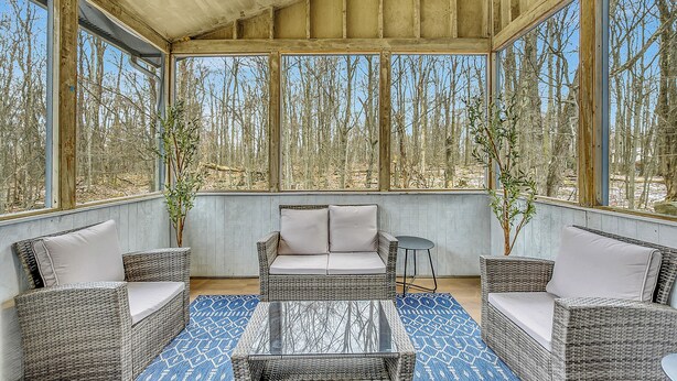 Un patio cubierto con mobiliario de mimbre, una mesa de vidrio y ventanas amplias que dan a una vista del bosque.