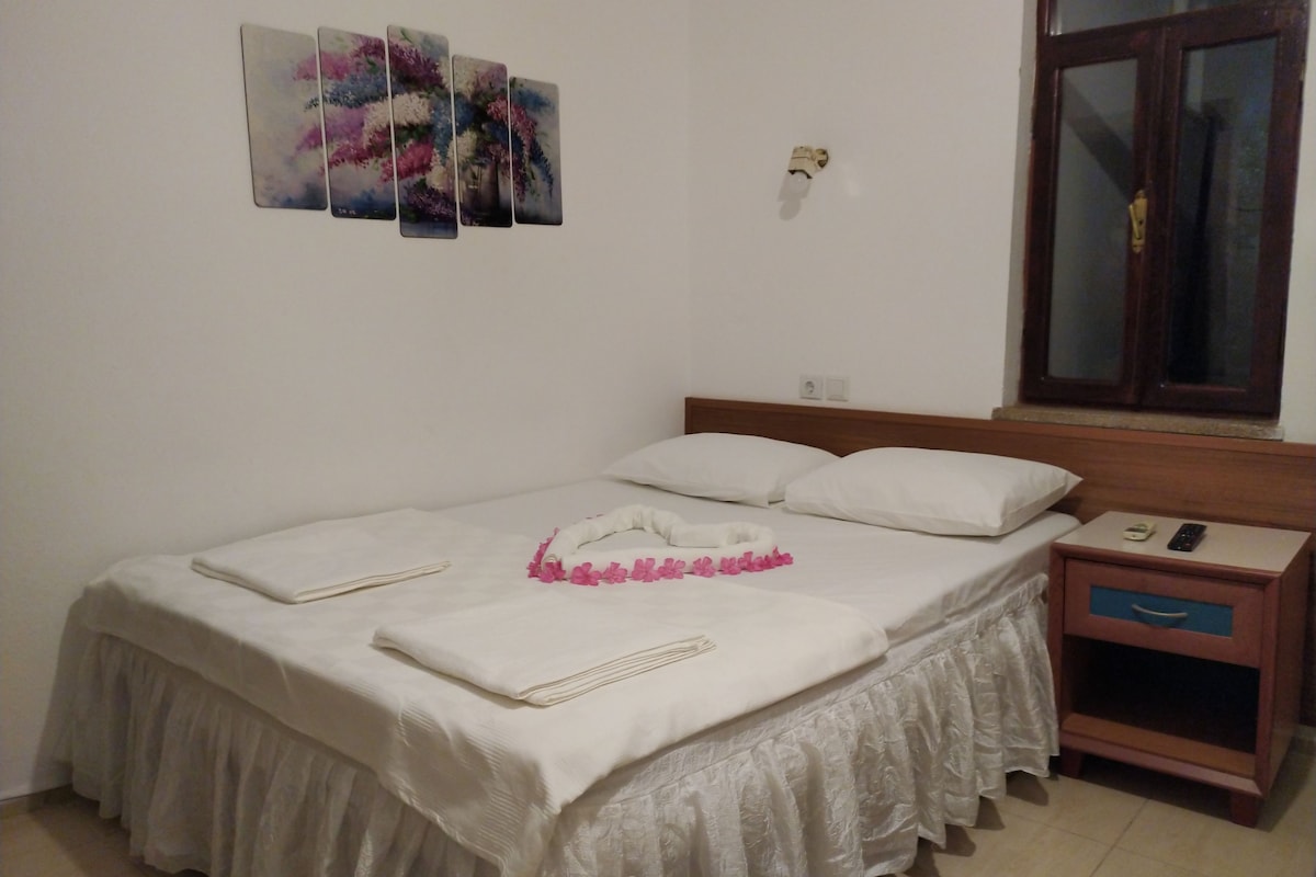 A neatly made bed with a white duvet and pillows, a bedside table with a remote control, and a window with a wooden frame.