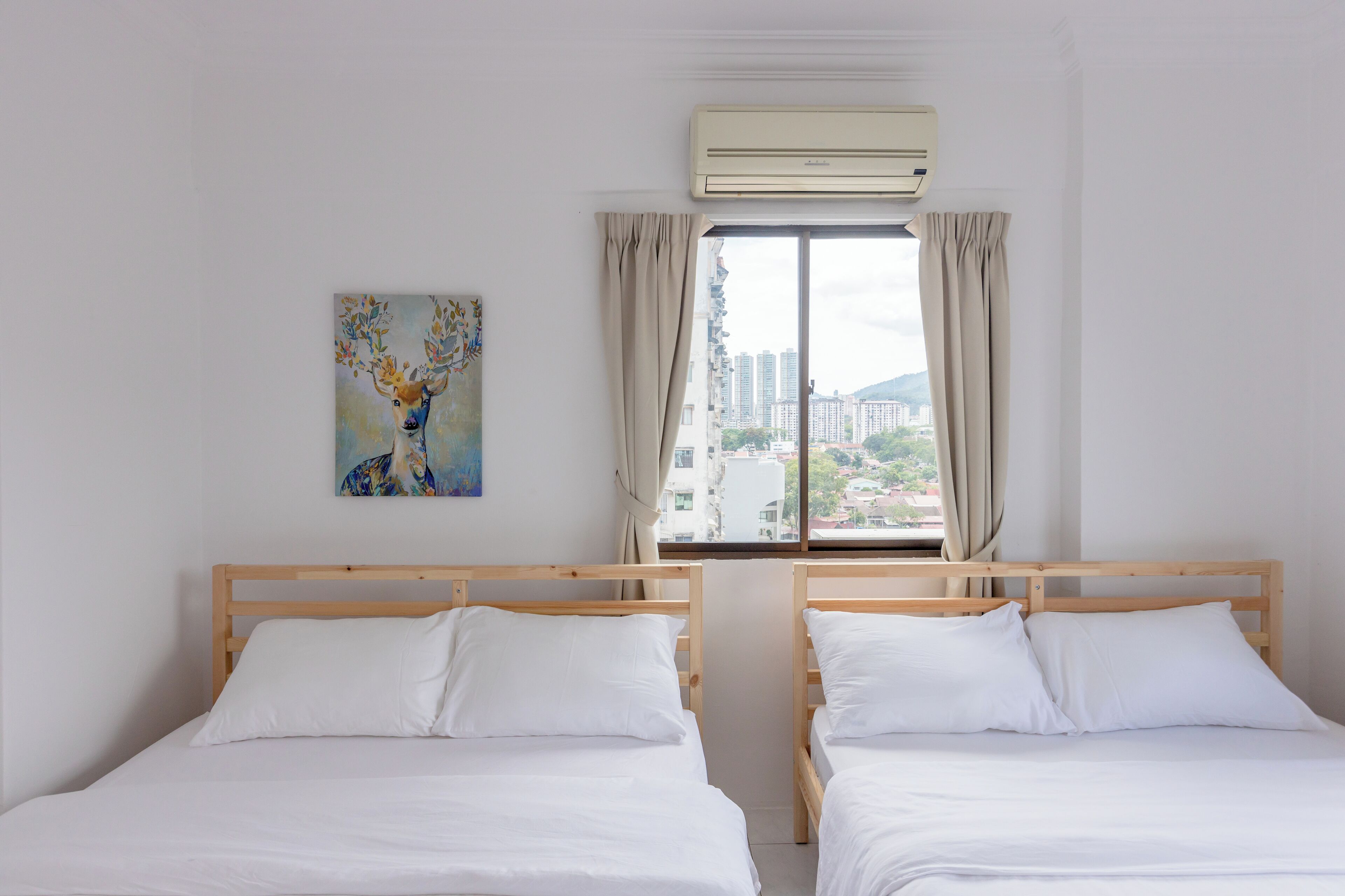 A hotel room with two wooden beds, white bedding, a window with curtains, an air conditioner, and a painting on the wall.