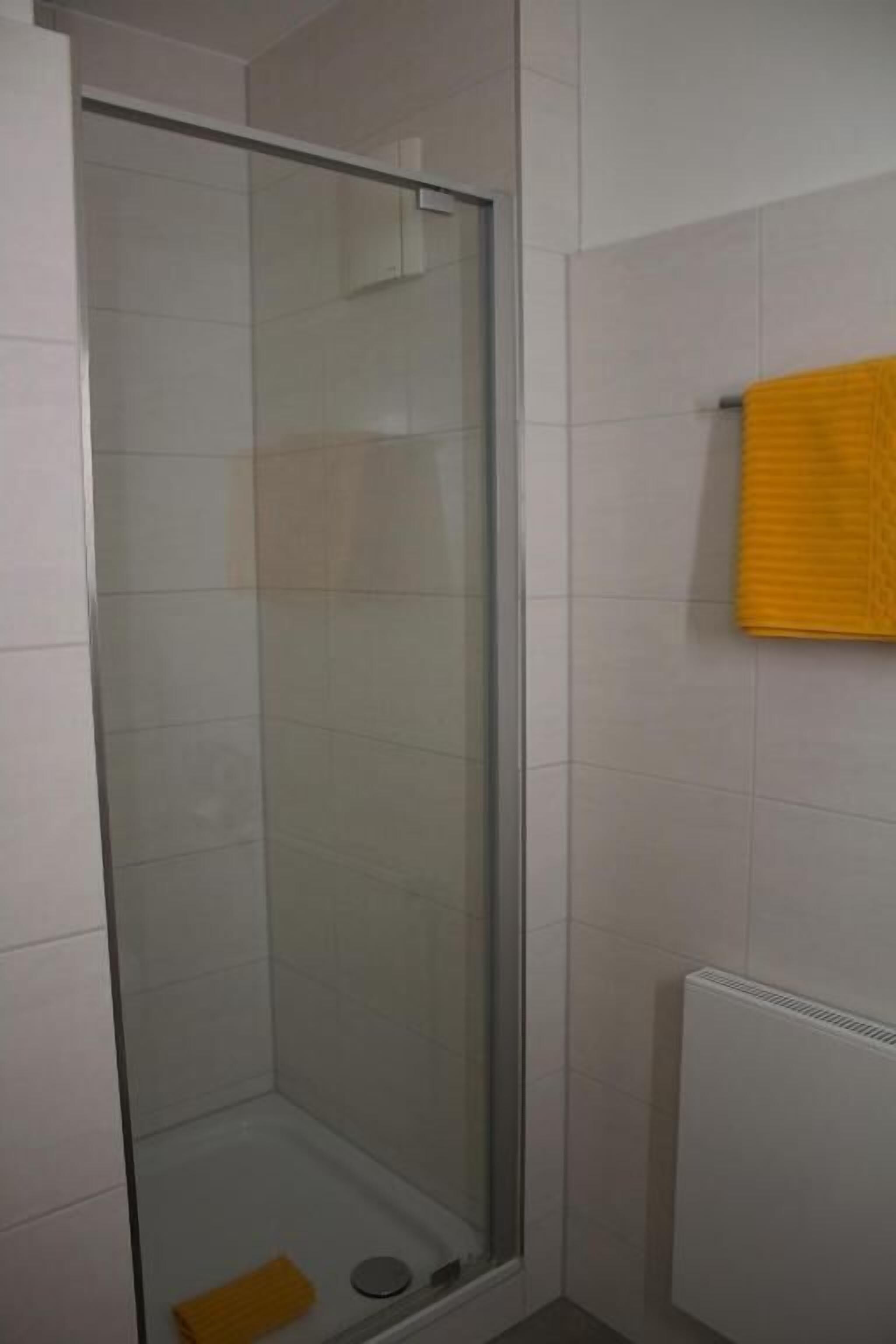 A shower with a glass door, white tiled walls, and a yellow towel hanging on the wall.