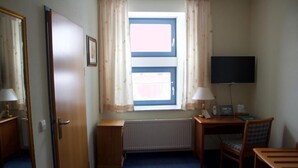 Une petite pièce bien éclairée, avec une seule fenêtre, un bureau avec une chaise, un téléviseur et un radiateur.