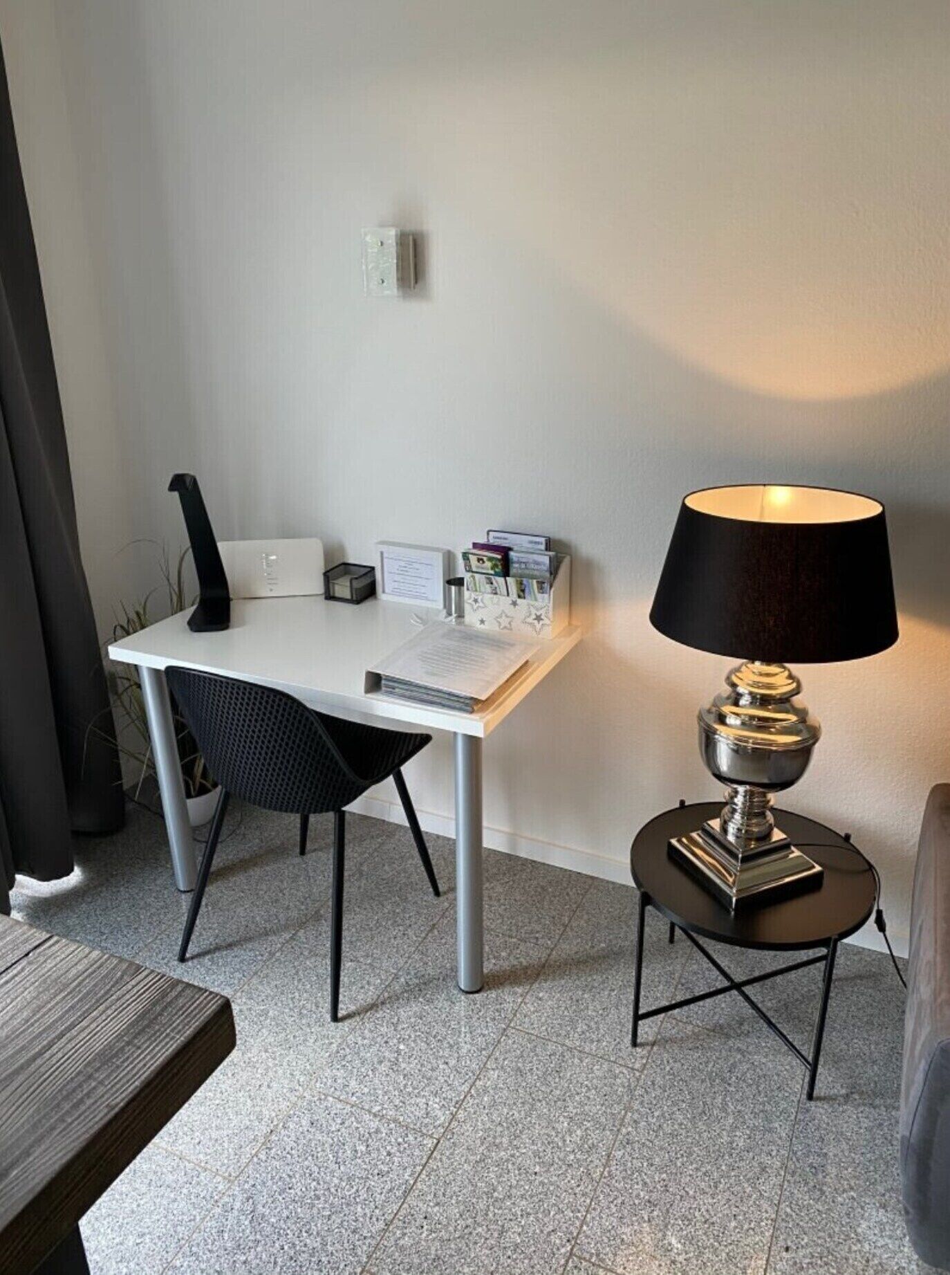 A small, modern room with a white desk, a black chair, a lamp, and a small table.