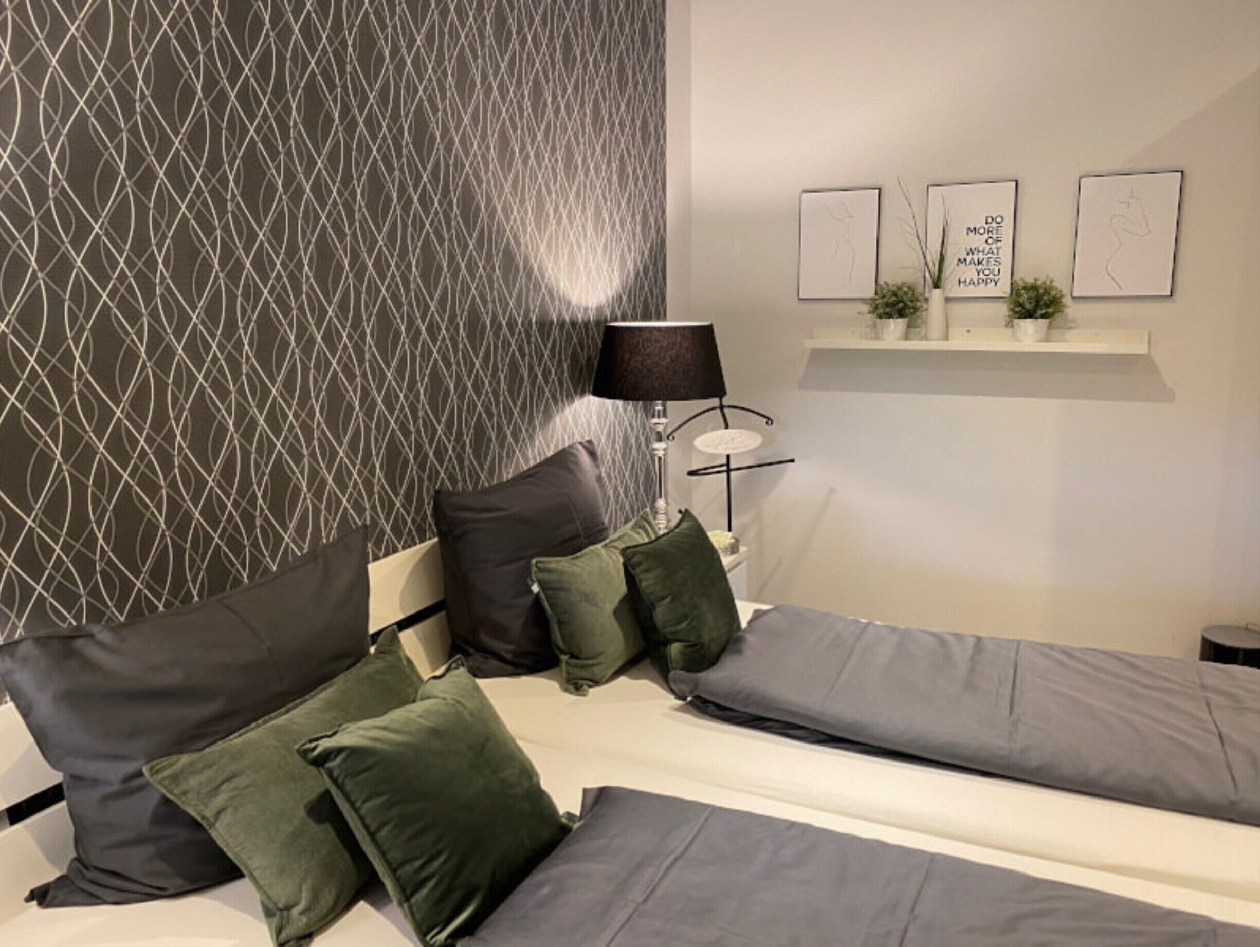 Ein Schlafzimmer mit strukturiertem Wandpapier, einem Nachttisch mit Lampe und drei gerahmten Motivationssprüchen an der Wand.