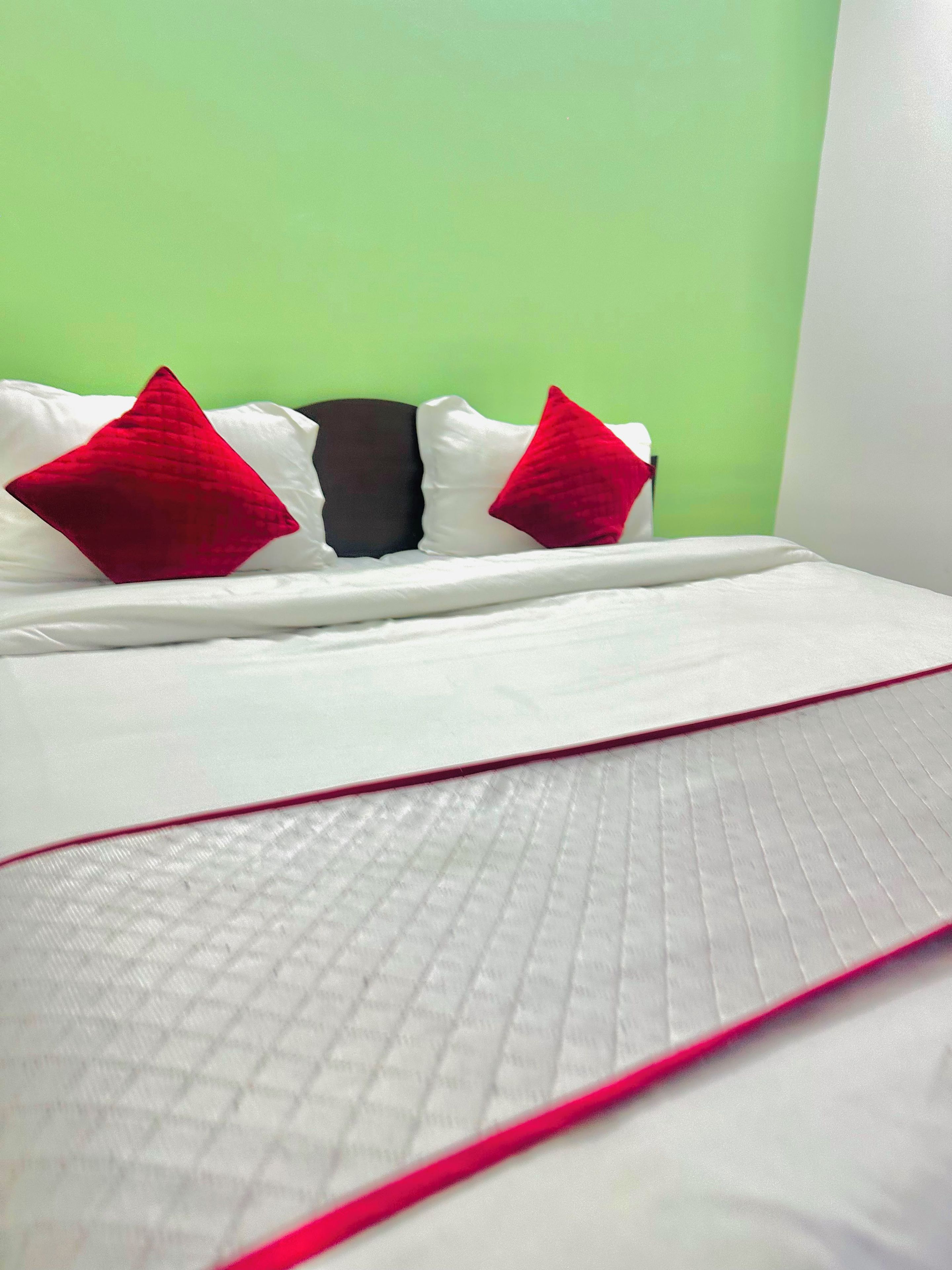 Una cama con ropa de cama blanca y dos almohadas rojas, una pared verde, y una almohada blanca con una almohada negra detrás.