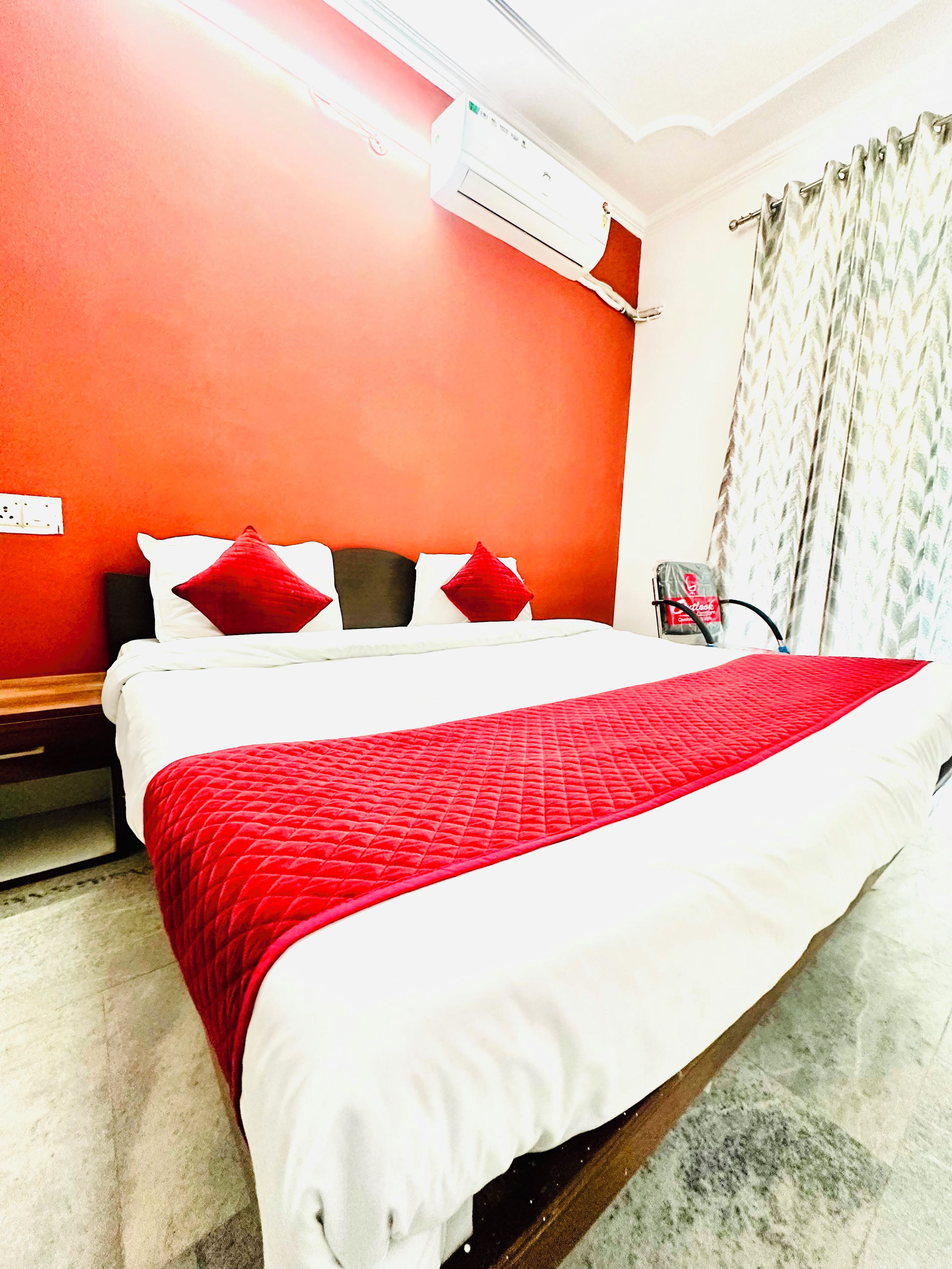 A hotel room with a bed, a red and white bedspread, a white wall, and a window with curtains.