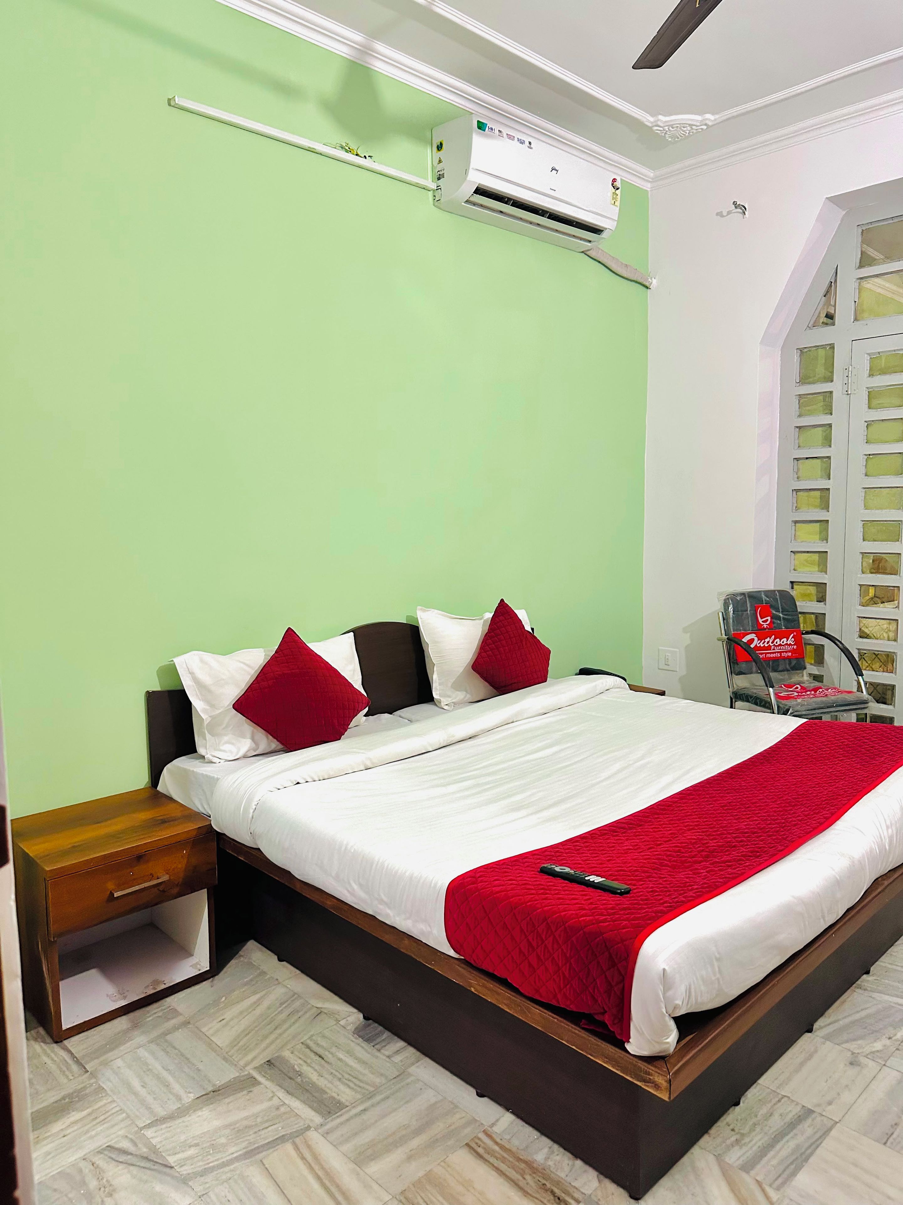 A neatly made bed with a red blanket, a wooden nightstand with a drawer, and an air conditioning unit mounted on the wall.
