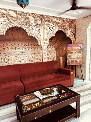 A room with a red sofa, a coffee table with various items, and a wall with intricate patterns. - Angel paradise (Jaipur)