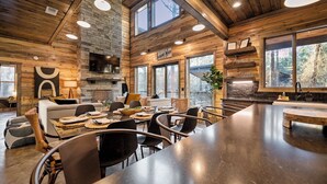A spacious wooden interior with a long dining table, comfortable seating, a fireplace, and large windows offering views of the outdoors. - 4BR Broken Bow Cabin Sleeps 12 | Hot Tub, Arcade, Firepit (Broken Bow)