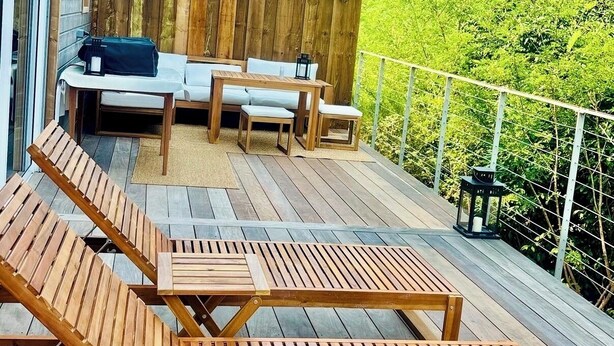 Eine Holzterrasse mit Liegestühlen und einem kleinen Tisch, ein Holzzaun und üppiges Grün im Hintergrund.
