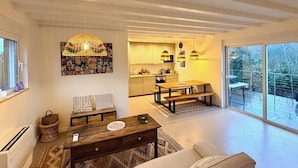 Ein modernes Wohnzimmer mit Couch, Esstisch und einer Bank. Im Hintergrund befindet sich eine Küche und ein großes Schiebeglastür, das auf einen Balkon führt.