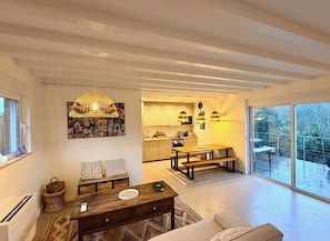 A modern living room with a wooden coffee table, a sofa, and a dining area with a bench. There is a kitchen in the background and a large sliding glass door leading to a balcony. - Charming new lodge house on the edge of a forest, in a natural setting close to the ocean. (Ondres)