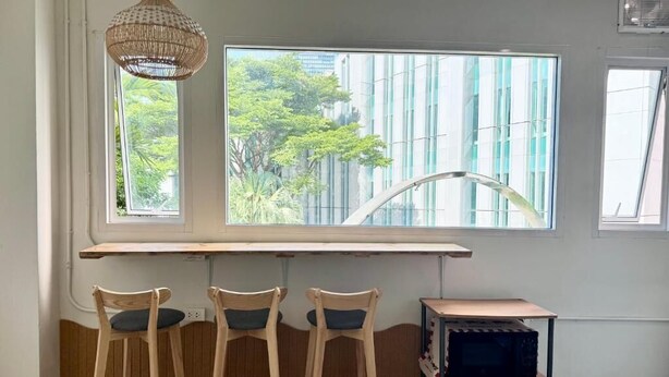 A room with a large window, a wooden table, three chairs, a hanging light fixture, and a small shelf with items. - Nana Best Hostel (Bangkok)