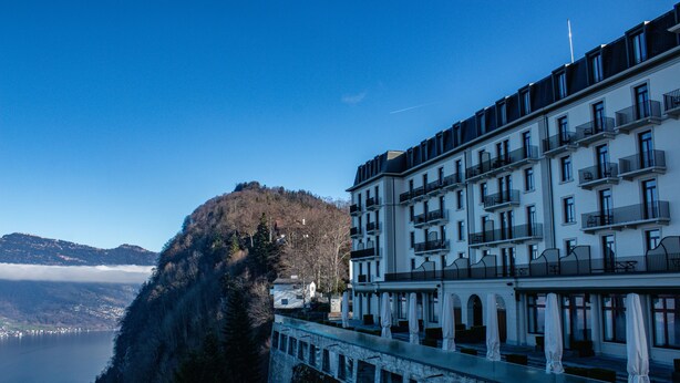 A hotel with a terrace overlooking a lake, a mountain in the background, and a clear sky.