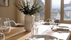A dining table set with plates, glasses, and cutlery, a vase with green foliage, and a window with a view of buildings.