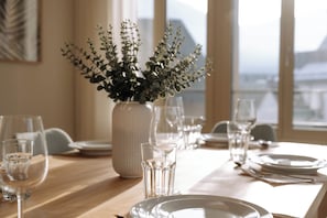 Une table à manger dressée avec des assiettes, des verres et des couverts, un vase rempli de feuillage vert, et une fenêtre donnant sur des bâtiments.