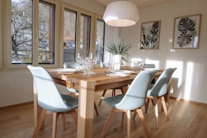 Un coin repas avec une table en bois dressée pour six personnes, entourée de six chaises, et de grandes fenêtres offrant une vue sur les arbres et les bâtiments environnants.