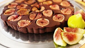 Una tarta de chocolate con higos en rodajas sobre un plato de metal, acompañada de higos enteros y una manzana verde sobre una mesa de madera.