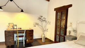 Un dormitorio con cama, escritorio y silla, jarra con flores, ventana con cortinas y lámpara en una mesita de noche.
