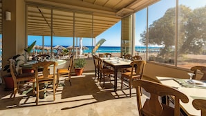 Un comedor iluminado por el sol con vista a la playa, muebles de madera y un ventanal con vistas al océano.