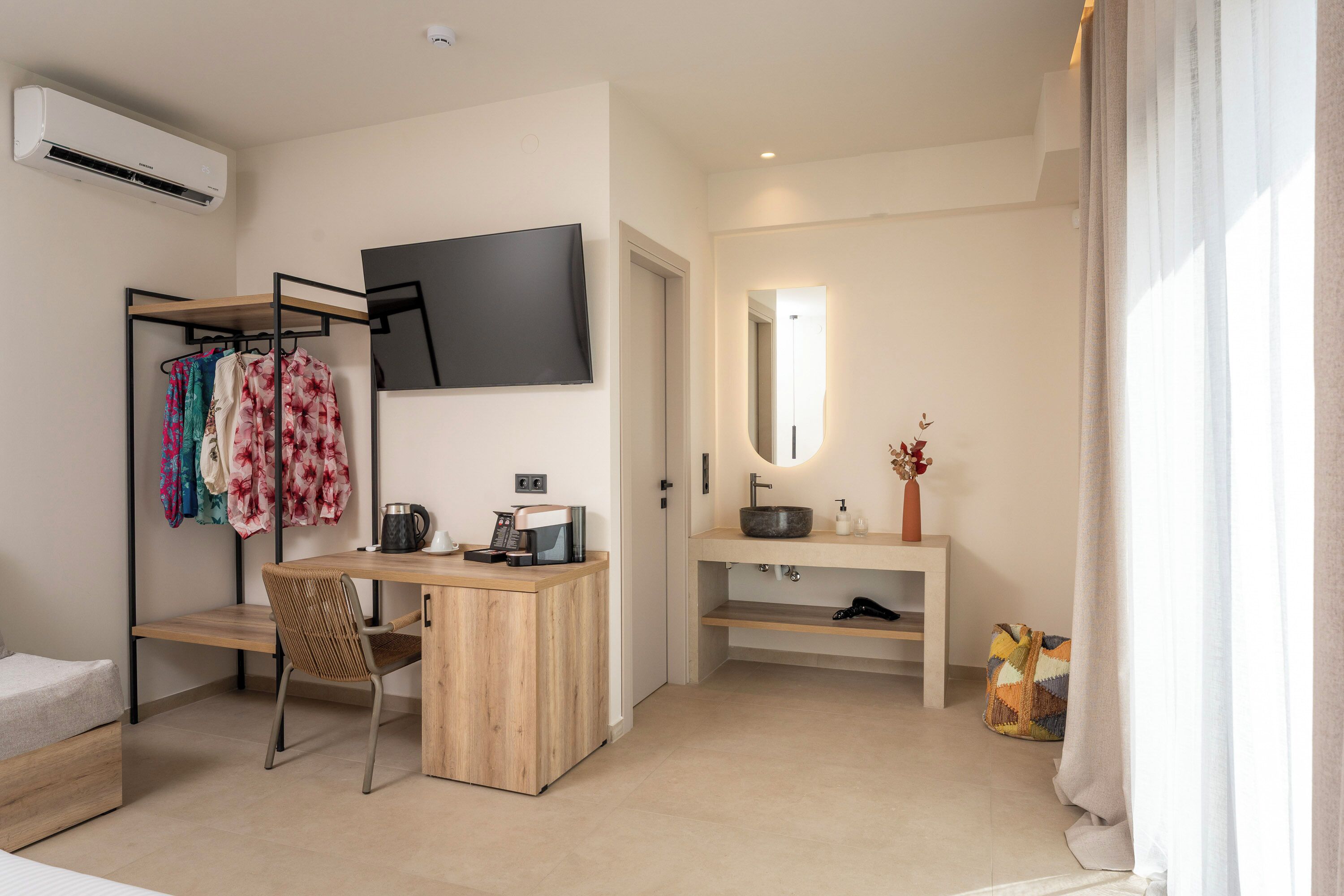 Una habitación de hotel moderna con un escritorio de madera, un televisor de pantalla plana, un armario con ropa, un baño con lavamanos y espejo, y una ventana con cortinas.
