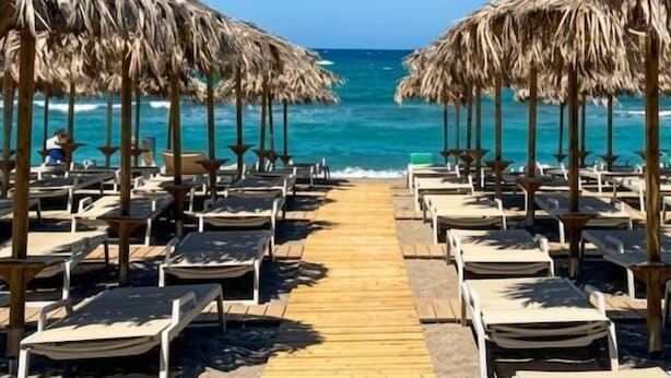 Un paseo marítimo frente a la playa que conduce a sillas de descanso bajo sombrillas de paja, con el océano al fondo.