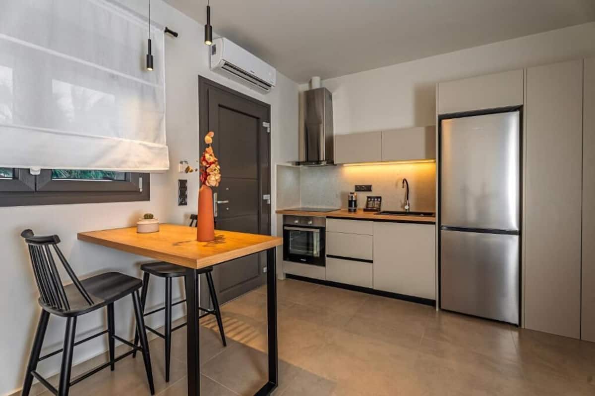 A modern kitchen with a wooden table, two black bar stools, a stainless steel refrigerator, and a built-in oven.