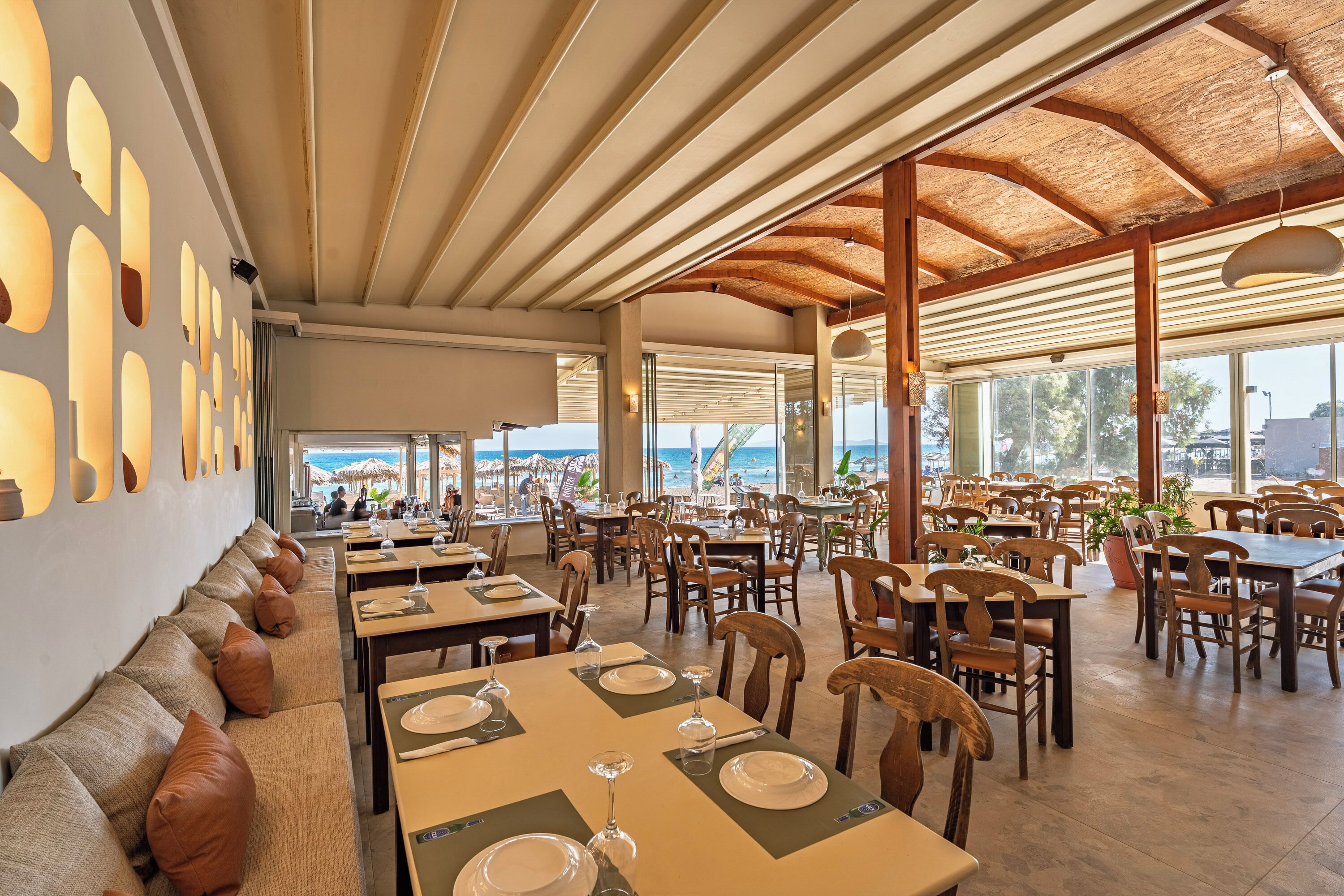 A spacious restaurant with high ceilings, wooden beams, and large windows offering a view of the beach.