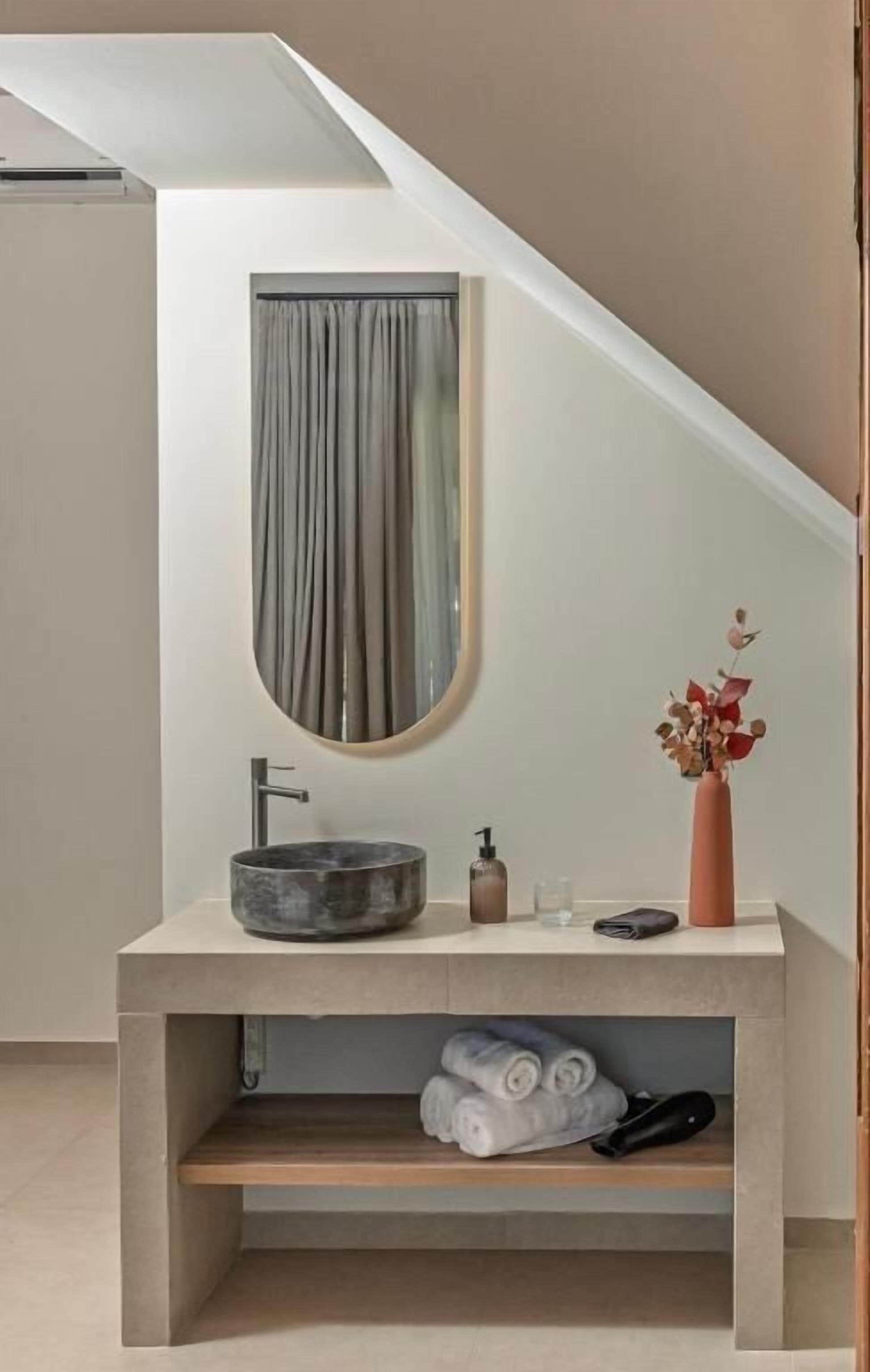 Un baño moderno con un lavabo redondo, un espejo y un jarrón con flores secas. Hay toallas y un bolso negro en un estante debajo del lavabo.