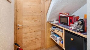 Ein kleiner, abgeschlossener Raum mit einer Holztür, einem Regal voller Gegenstände, einer Mikrowelle und einer Kaffeemaschine.