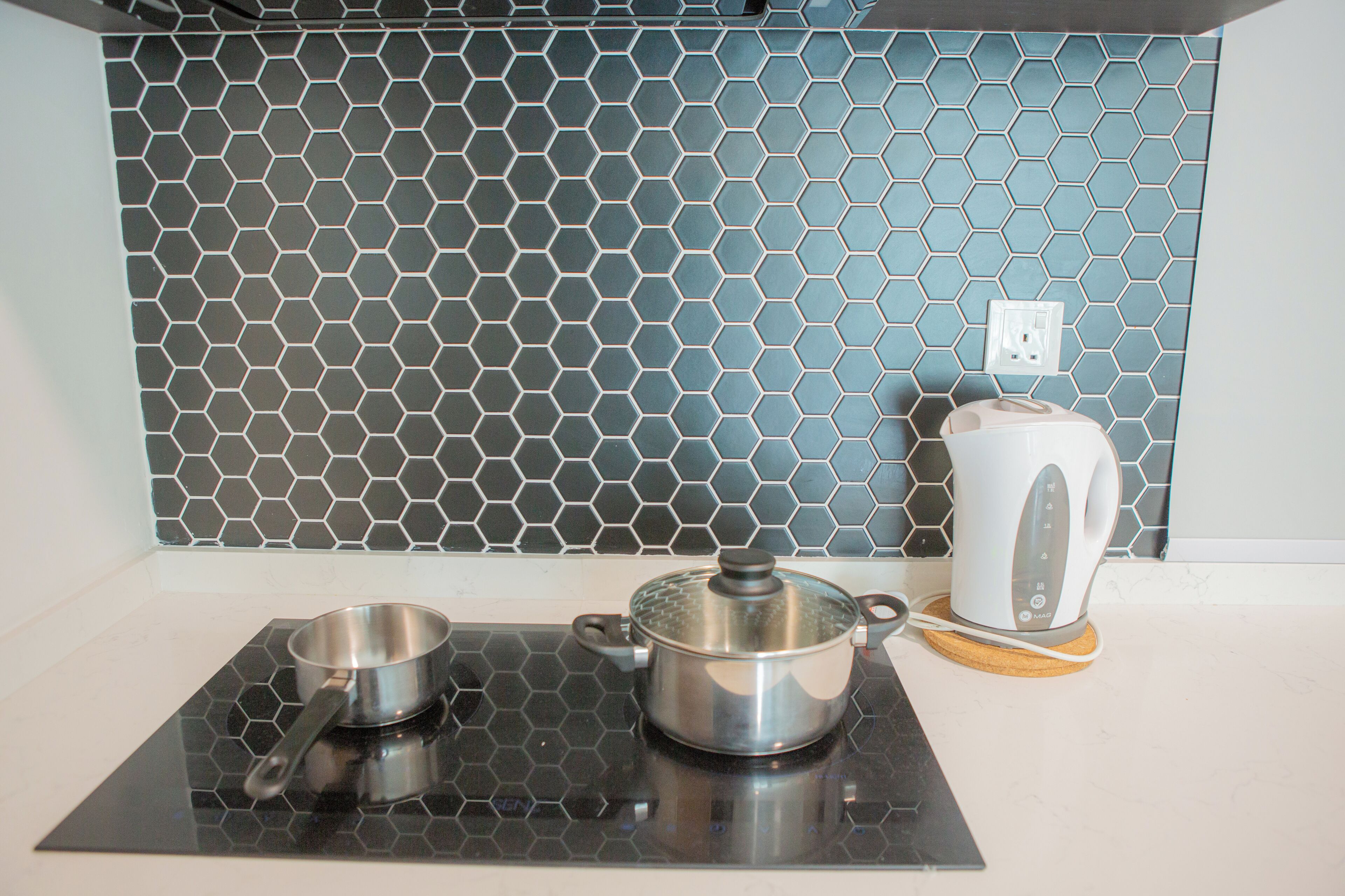A kitchen with a black hexagonal backsplash, a stainless steel pot on an induction cooktop, and a white electric kettle on a coaster.