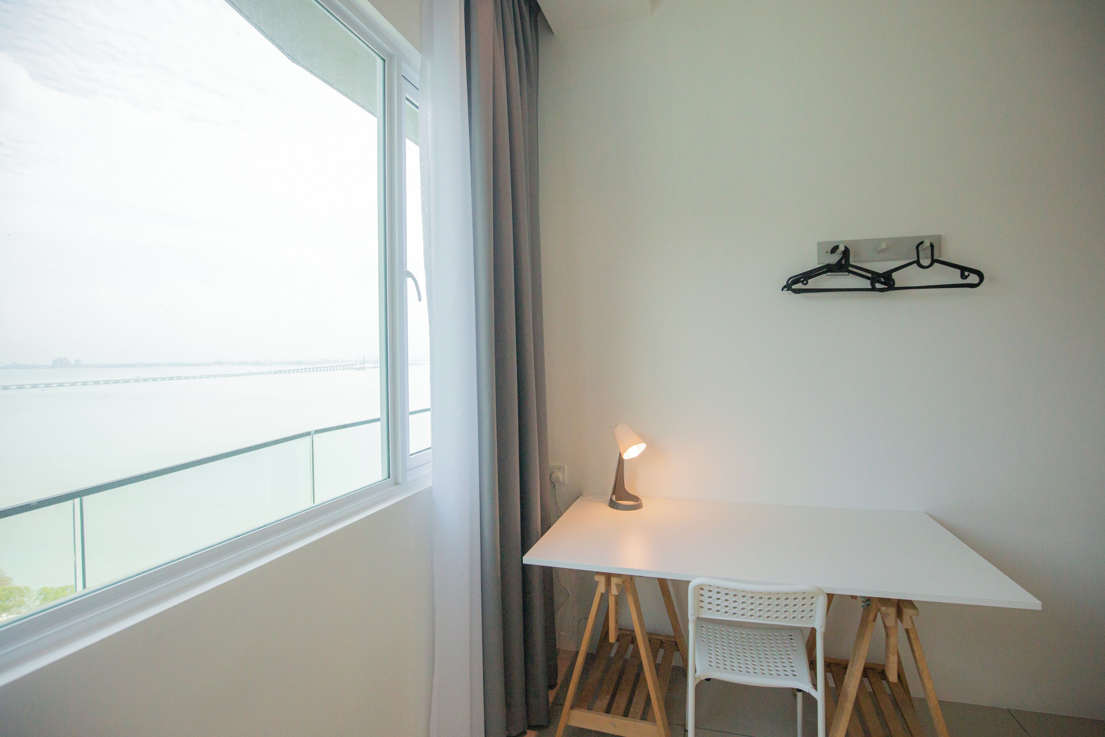 A room with a white table, a lamp, a chair, a window with curtains, and a wall-mounted hanger.