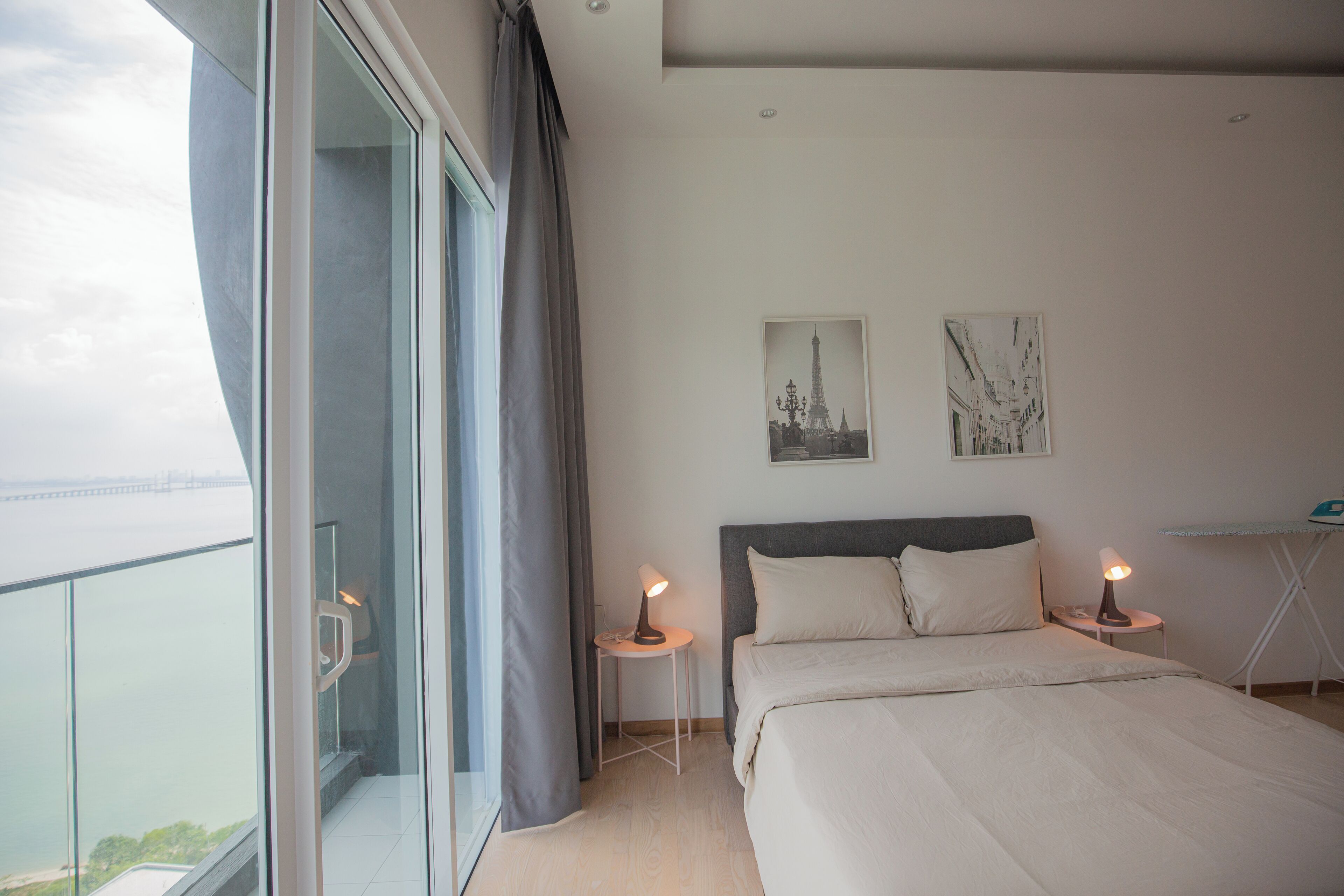 A modern bedroom with a large bed, two bedside tables with lamps, a view of the city, and artwork on the wall.