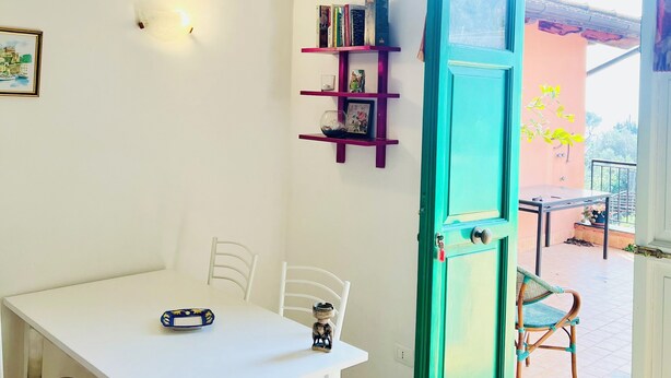 Une pièce avec une table blanche, des chaises, une porte de couleur turquoise, une bibliothèque et une vue sur l’extérieur.