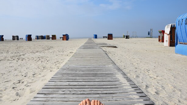 Ein hölzerner Steg führt zu Strandhäusern an einem Sandstrand.