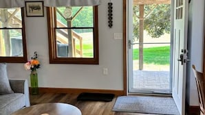 Une pièce avec un plancher en bois, un canapé gris, une table ronde, une porte donnant sur l’extérieur et une fenêtre avec des rideaux.