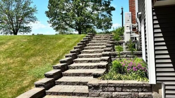 Un escalier en pierre mène à un bâtiment doté d’une cheminée en briques, qui est entouré de pelouse et d’arbres.