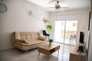 Una sala de estar con un sofá color beige, una mesa de centro de madera, un televisor y un ventilador de techo.