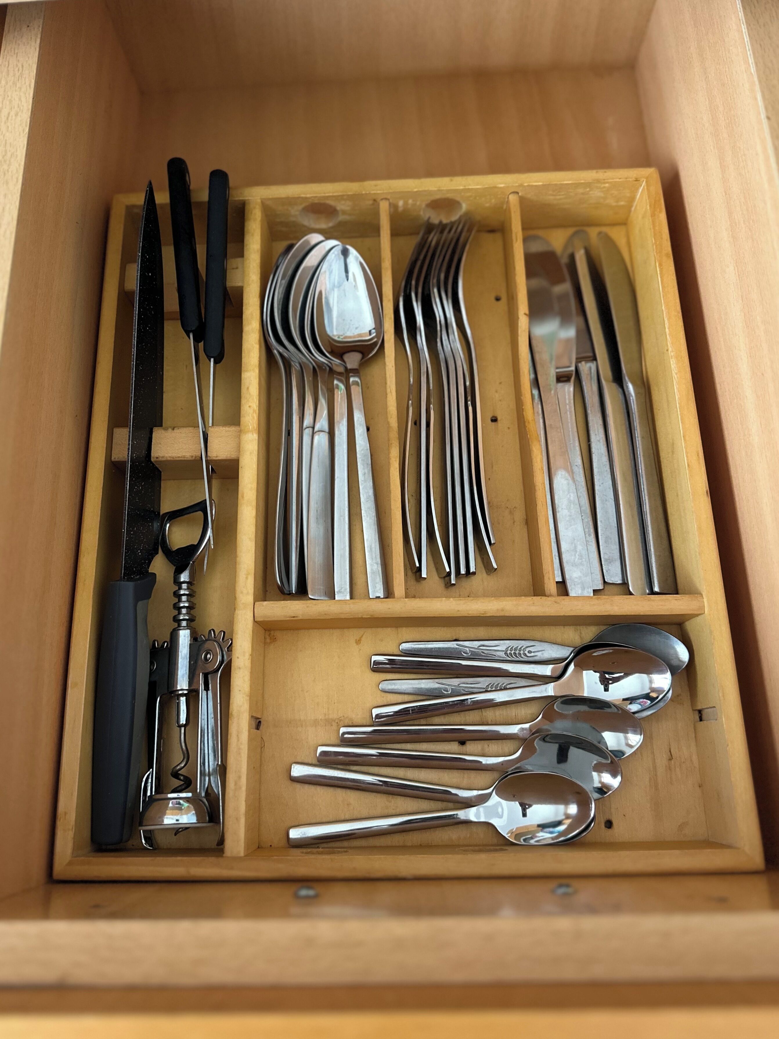 Un cajón de madera con varios utensilios de cocina ordenados, incluyendo cuchillos, cucharas, tenedores y un sacacorchos.
