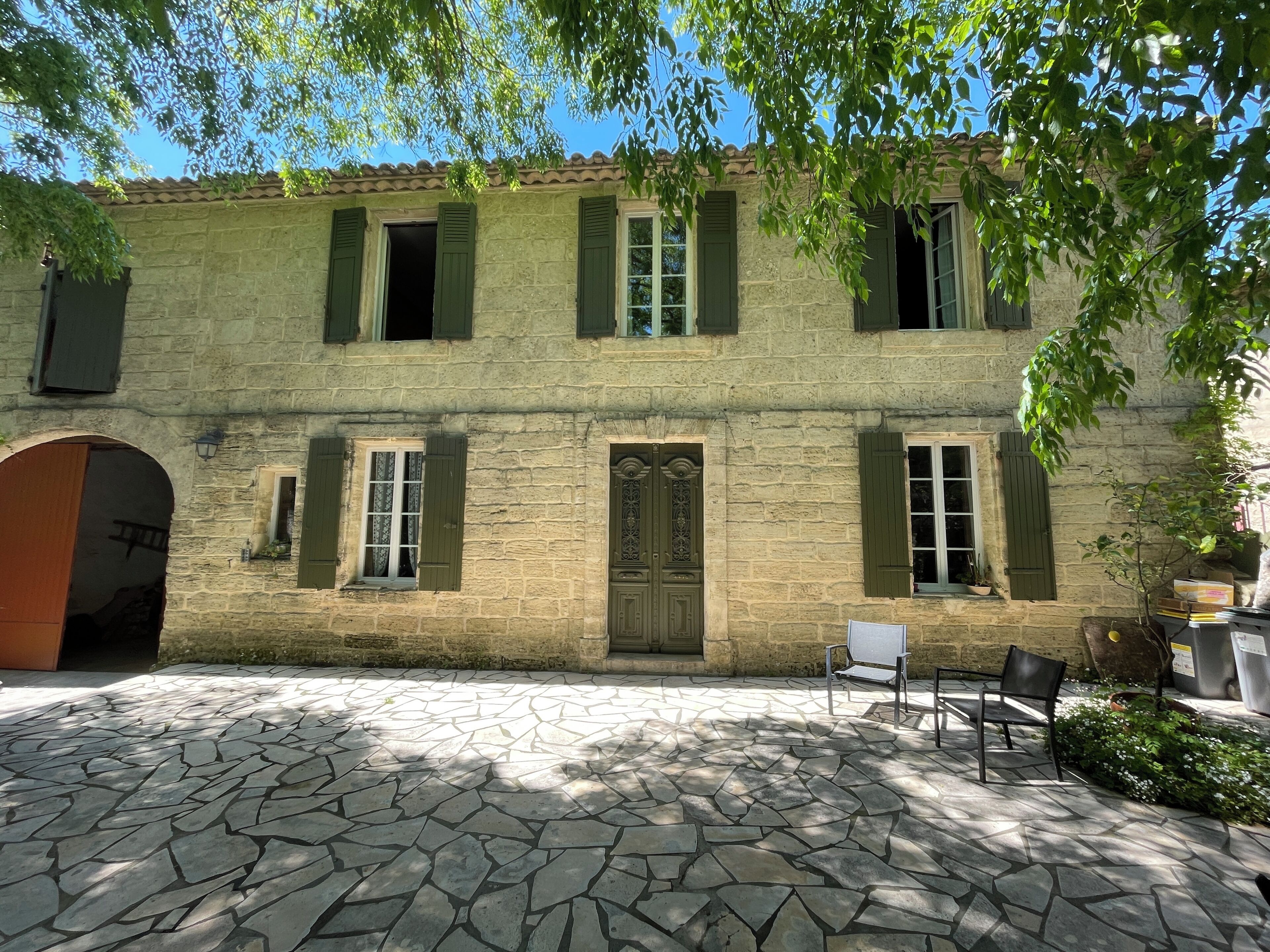 Un edificio de piedra con contraventanas verdes, una puerta central y un patio con suelo de piedra y mobiliario exterior.