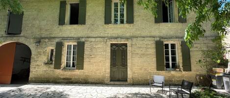 A stone building with green shutters, a central door, and a courtyard with stone flooring and outdoor seating.