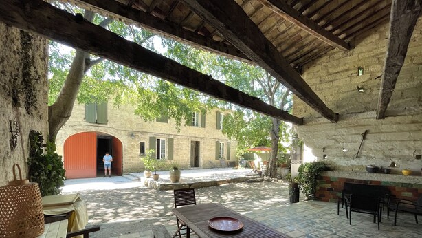 An outdoor patio area with stone flooring, wooden tables and benches, and a view of a building with a red archway. - 19th-century farmhouse combining authenticity and comfort (Gallargues-le-Montueux)