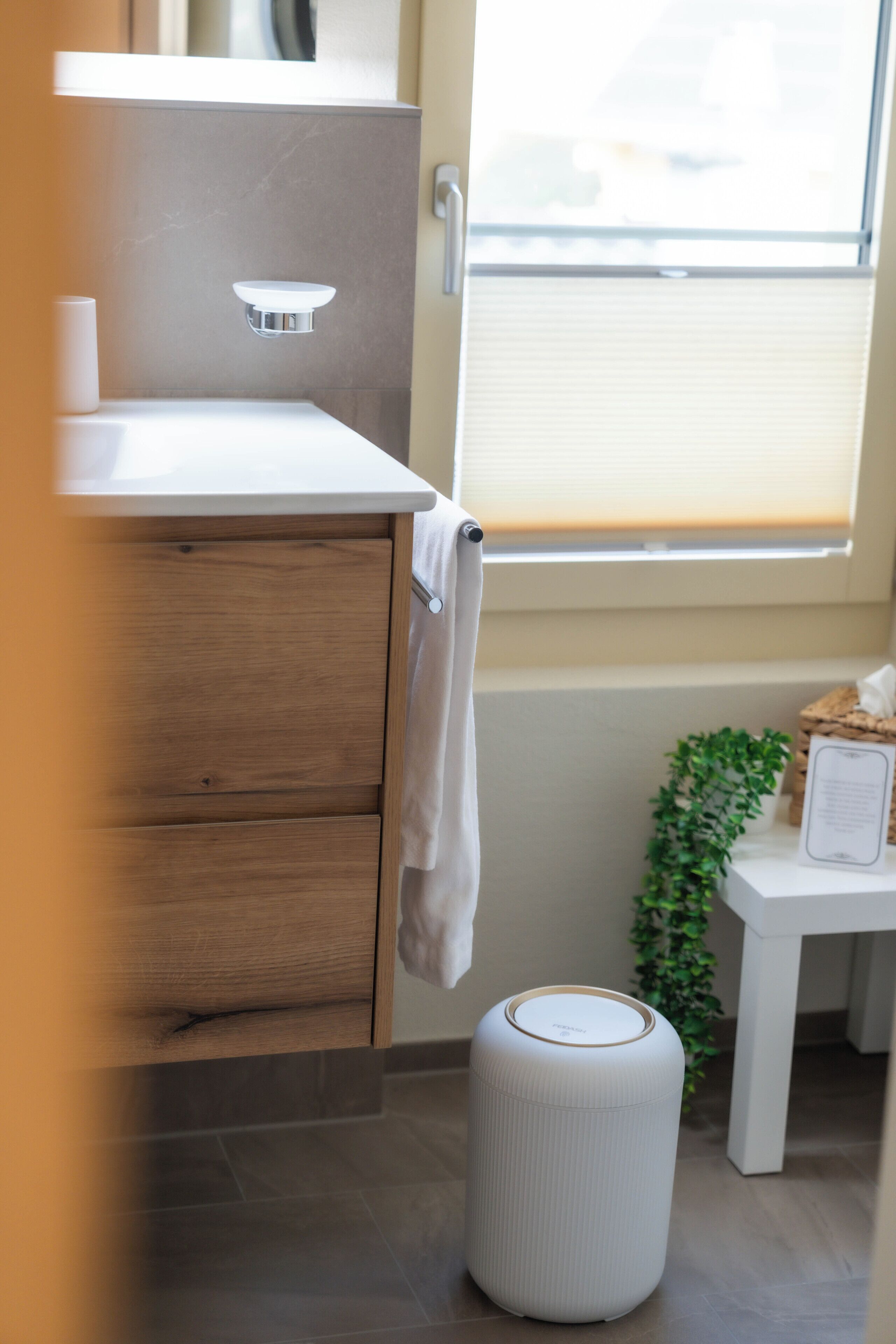 A bathroom with a wooden vanity, a white sink, a towel hanging on the door, a small trash bin, and a window with blinds.