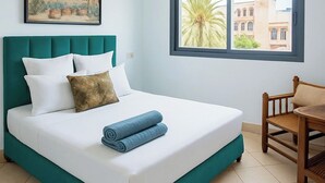Un letto ben rifatto con lenzuola bianche e biancheria verde acqua, una sedia di legno e una finestra con vista su edifici e una palma.