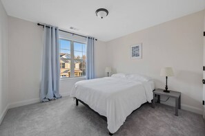 Een slaapkamer met een groot raam, een bed, een nachtkastje met een lamp, en een plafondlamp.