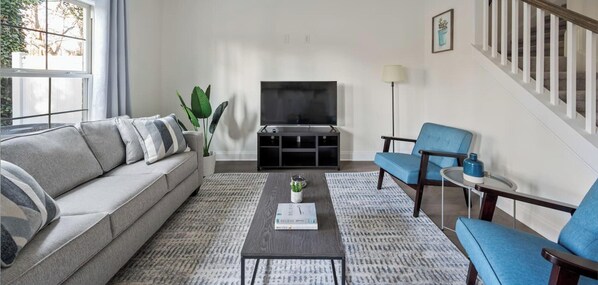 Ein Wohnzimmer mit Sofa, Couchtisch, Sesseln, einem Fernseher und einer Treppe.