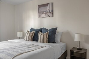 Ein ordentlich gemachtes Bett mit weißer Decke und mehreren Kissen, ein Nachttisch mit einer Lampe und ein an der Wand hängendes Bild von einem Balkon.