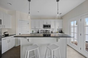 Una cocina moderna con armarios blancos, una isla central con taburetes, electrodomésticos de acero inoxidable e iluminación colgante.
