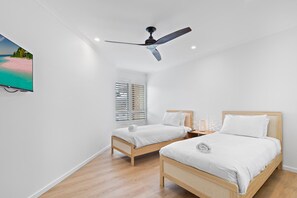 Ein Schlafzimmer mit zwei Einzelbetten, einem Deckenventilator, einem an der Wand montierten Fernseher und einem Fenster mit Jalousien.