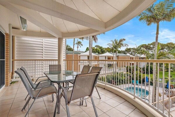 A balcony with a glass table and chairs, overlooking a swimming pool and palm trees. - Macquarie Lodge Apartments (Sunshine Coast)