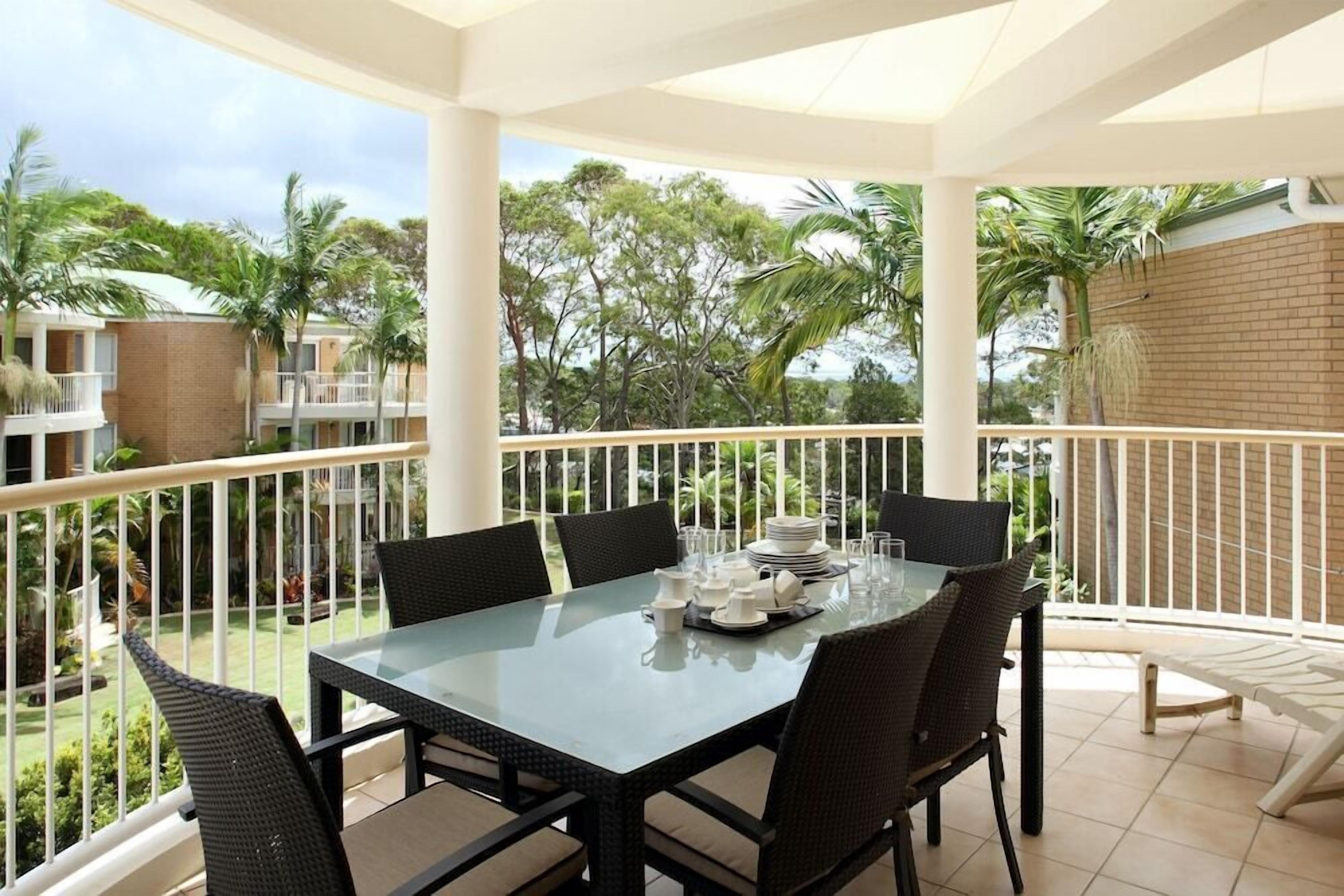 A balcony with a glass table set for dining, surrounded by wicker chairs, overlooking a garden with palm trees and a brick wall.